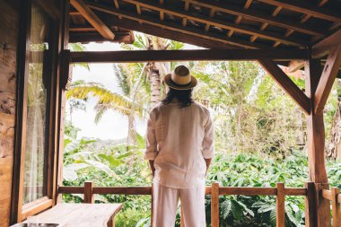 Beyaz elbiseli ve hasır şapkalı isimsiz bir adamın arka plan görüntüsü gür tropik ağaçlara ve bitkilere karşı ahşap verandada duruyor.