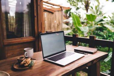 Gündüz aydınlatma tuş takımı paneli ile boş ekranlı modern ahşap masa ve yanında not defteri ve kalem varken tepside servis edilen kahve kruvasanı.