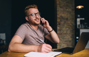 Görsel koruma için optik gözlük takan yetenekli bir erkek blogcu cep telefonu uygulamasını kullanarak birlikte çalışma alanında telefon kullanırken, bilgisayar ve ders kitabı ile arayan beyaz bir adam.