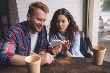 Kafede dinlenirken, bir fincan kahve ve cep telefonuyla ahşap masada oturan, gündelik kıyafetli, pozitif genç bir çift.