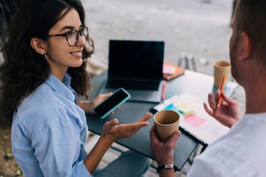 Akıllı telefonlu genç bir bayanın yan görüntüsü dizüstü bilgisayar ve pano ile masada otururken açık kafede erkek meslektaşıyla birlikte çalışırken ve belirsiz el hareketleriyle birbirlerine bakarken.