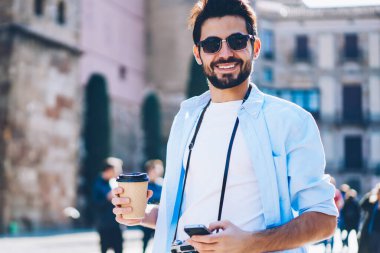 Klasik fotoğraf makinesi ve kahveyle gezen mutlu sakallı bir turist. Mimari şehrin kentsel ortamında yürüyor ve kameraya gülümsüyor. Elinde telefonla gezen güneş gözlüklü genç adam.