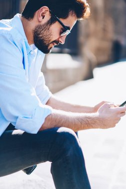 Yan görünüm casual güneş gözlükleri smartphone köknar yayın şehir merkezinde oturan Web sitesinde metin yazarak erkek blogger giymiş. Genç adam 4 Internet bağlantısı üzerinden çevrimiçi arkadaşlarınızla sohbet