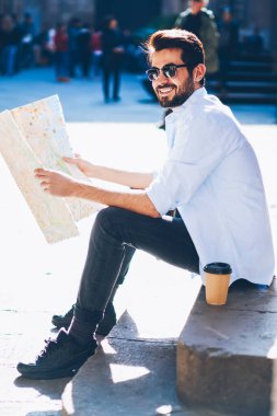 Güneş gözlüklü neşeli sakallı turist elinde seyahat haritası tutarken gülüyor ve şehir merkezindeki eski inşaatın güzelliğinin tadını çıkarıyor yaz gezisi sırasında merdivenlerde oturuyor.