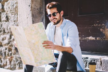 İlginç yerleri olan mutlu stil sahibi genç adam harita okuyor ve şehrin aşağısındaki mimari merdivenlerde otururken rotasını belirliyor. Olumlu sakallı turistler tatil beldelerini arıyor.