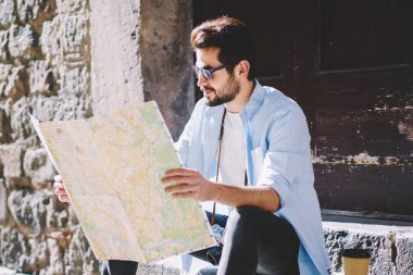 Düşünceli sakallı genç turist günlük kıyafetlerini giymiş seyahat haritasını dikkatle okuyor ve şehir merkezindeki merdivenlerde otururken bir sonraki gösteri için uygun rotayı belirliyor.