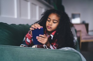 Serene Afro-Amerikan kızı, 6-8 yaşlarında, yeşil bir kanepede uzanmış, akıllı telefonuna dalmış. Kırmızı kareli gömlek ve mavi kot pantolon giyiyor. Merakını ve dikkatini gösteriyor. Ev ortamında.