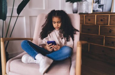 Uzun saçlı, rahat bir oturma odasında dinlenirken koltukta oturan ve akıllı telefonları karıştıran sıradan giysili Afro-Amerikan bir çocuk.