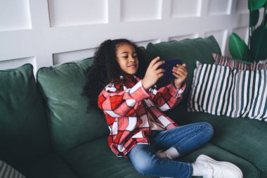 Rahat bir oturma odasında dinlenirken kanepede yastıklarla oturan ve akıllı telefondan oyun oynayan, gündelik kıyafetli genç Afrikalı Amerikalı kızın yüksek açısı.