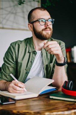 Gözlük Genç adam portresi bir şey notlar ve kameraya, seyir defter, gelecekteki proje hakkında düşünme ciddi profesyonel mimar için ev ödevi yazma gözlük öğrenci sakallı