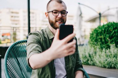 Sokak kafe masada oturan ve yeni blog için video yapmak için smartphone kullanarak hipster adam gülümseyen hareket eden telefon tutarak ve modern ön kamera ile selfie fotoğraf çekmek yakışıklı sakallı erkek