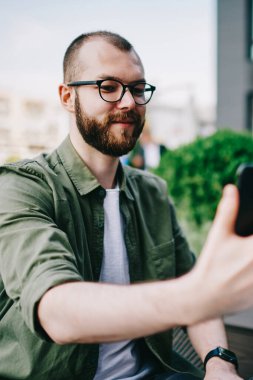 Trendy erkek holding cep telefonu cep telefonu kamera ve kendini açık havada kafede oturan ve selfie fotoğrafı tıklatın smartphone kullanarak beyaz hipster adam alarak fotoğraf gülümseyen elinde sakallı