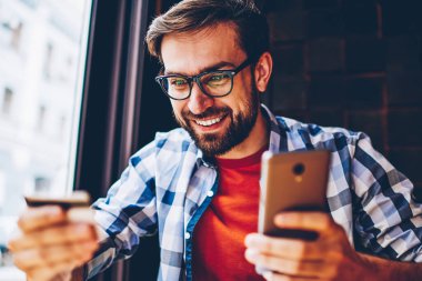 Smartphone cep telefonu ile internet bağlantısı ile internet sitesinde ödeme yapmadan elinde banka kartı ile neşeli sakallı öğrenci. Kartvizit para hareket için telefon tutarak bakarak olumlu genç adam