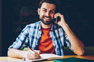 Smartphone üzerinde arama ve notlar not defteri alanı coworking oturan yapım sırasında kameraya gülümseyen başarılı sakallı öğrenci portresi. Pozitif hipster adam telefon aygıtı üzerinde iletişim