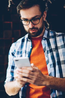 Dalgın sakallı hippi blogger gelen bildirim okuma ve ağlar Smartphone cep telefonu ile sohbet bağlı 4 g için Internet. Genç adam cep telefonu online video izlerken rahat gömlek giymiş