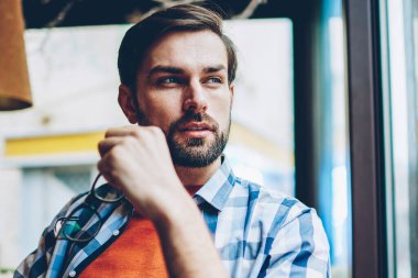 Optik gözlük pencereden dışarı bakıyor ve kendi başlangıç projesi geliştirmek için yaratıcı fikirler üzerinde düşünme sırasında elinde tutan rüya gibi yakışıklı genç adam. Düşünceli hipster adam planları üzerinde durulması