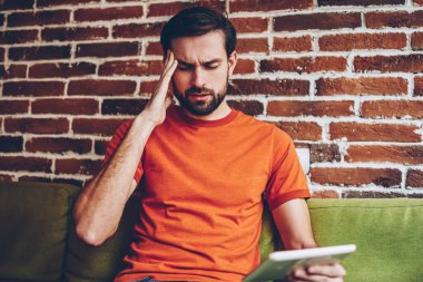 Turuncu gömlek giymiş stresli sakallı adam migren vardır. Mutsuz hipster adam alan coworking içinde modern tablet cihaz ile kanepede otururken baş ağrısı vardır. Dokunmatik yüzey ile yorgun erkek