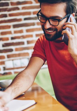 Neşeli smartphone aygıt üzerinde arama ve metin bilgileri dizüstü cihaz alan coworking içinde oturan Not Defteri'nde yazma genç adam sakallı. Telefonla konuşurken ve notlar olumlu hipster adam