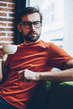 Dalmış genç adam lezzetli kahve elinde tutan ve pencereden dışarı bakıyor 20 yaşında sakallı. Düşünceli hipster adam gözlük vizyon düzeltme kafede oturan içecek zevk için