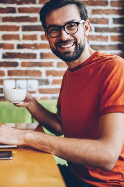 Portre kahve elinde tutan ve modern dizüstü bilgisayar yazılımı güncelleştirmek için 4 g bağlıyken kameraya gülümseyen başarılı sakallı genç adam Freelancer internet kafede oturan