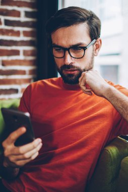 Sosyal ağlar üzerinde modern smartphone aygıt 4 g internet bağlantısı üzerinden video izlerken sakallı hippi blogger yoğunlaşmıştır. Dalgın genç adam üstünde hareket eden telefon aygıt Web sitesinde haber okuma