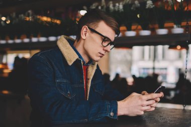 Dalgın hipster sohbet blogger online takipçileri smartphone ile bağlı 4 g internet kafede oturuyor. Kafede dinlenme telefon uygulama yükleme denim ceket genç adam