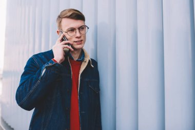 Smartphone üzerinde arama ve mobil faydaları işleci ile görüşmesi sırasında tartışırken rahat giyimli genç adam. Telefon ayakta duvarının yakınında kentsel ortamda konuşurken kot ceketli hipster çocuk