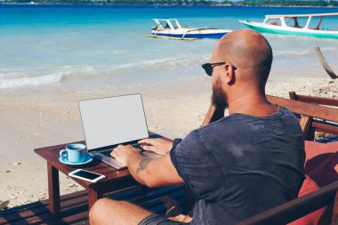 Arkadan görünüşü deniz kıyısında online profesyonel erkek serbest meslek sahibi netbook ile sahte kadar yaz yolculuk zevk ekran üzerinde klavye tatil sırasında çalışma dizüstü bilgisayar ile oturan genç adam
