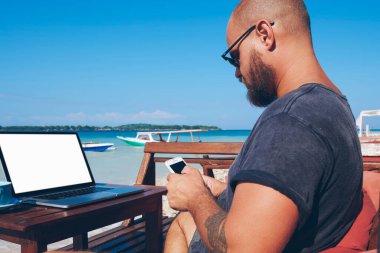 Modern teknolojiyi ve 4G interneti kullanan genç adam yazın okyanus sahillerinde uzaktan çalışıyor, milenyum erkek blogcu akıllı telefon uyarısını kontrol ediyor web sayfasının yolculuk içeriğini güncelliyor