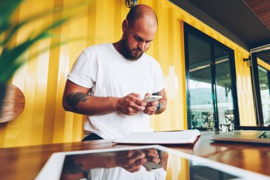 Ciddi hipster adam cep telefonu kısa mesaj, yeni uygulama ayakta masaüstü yakınındaki ofiste cihazları ile yüklemek için smartphone tutan genç bin yıllık erkek gönderme üzerinden sosyal ağlar sohbet