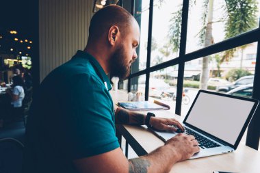Dizüstü bilgisayar yazarak metin için blog, yetenekli erkek serbest meslek sahibi mesafe işi netbook ile sahte ekran ve kablosuz internet üzerinden yayın ile kafeteryada oturan adam Genç sakallı