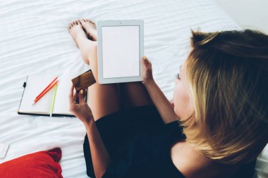 Genç hippi kız rahat bir yatakta dinleniyor taşınabilir bilgisayar ve çevrimiçi pişirme yoluyla alışveriş yapıyor, kadın evdeki dijital tableti kullanarak kredi kartından para kazanıyor.