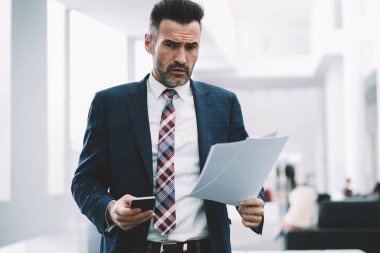 Deneyimli icra müdürü yarım uzunluk portre ofiste ayakta muhasebe raporları denetlerken kameraya bakarak formal giyim giymiş. Elinde telefon ile kendine güvenen gurur ceo