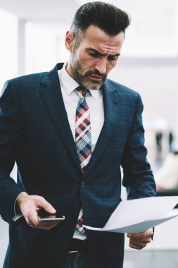 Dalgın ciddi girişimci dikkatle finansal raporlar ofiste ayakta okuma takım elbise giymiş. Olgun işadamı smartphone ile kağıt belgeleri kontrol elinde konsantre