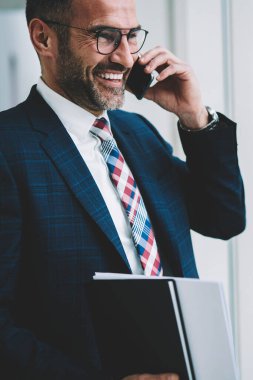 Pozitif olgun tüccar kıyafetli smartphone aygıt üzerinde iş ortağı ile telefon görüşmesi sırasında gülüyor. Cep üzerinde arama ve başarılı proje hakkında konuşmak neşeli gurur CEO'su orta yaşları