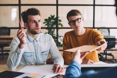 Ciddi bir grup genç erkek tasarımcı modern ofiste birlikte çalışıyor. Kafkasyalı bir adam, öndeki meslektaşlarına evrak veriyor. İşbirliği ve işbirliği kavramı.