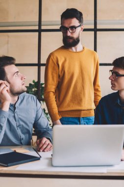 Dizüstü bilgisayar ve çevrimiçi uygulama işbirliği yaparak yeni bir proje için fikir alışverişinde bulunan yetenekli erkek meslektaşlar, ofiste çalışırken birbirleriyle konuşan bir grup genç.