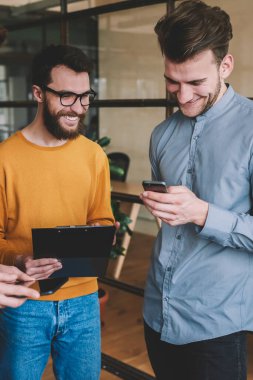 Başarılı erkek partnerler, sosyal ağda akıllı telefon cihazıyla komik fıkralar okumaktan zevk alıyor ve gülüyor, genç pozitif erkekler modern ofiste iletişim kuruyor ve teknolojiyi kullanıyorlar.