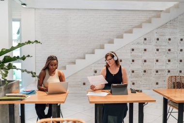 Modern bir çalışma alanında dizüstü bilgisayarlar ve belgelerle ilgili görevleri üzerinde çalışan iki odaklanmış kadın profesyonel, konsantrasyon ve modernite birleşimi