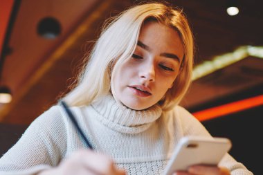 Zeki kız öğrenci ev ödevi yazma üzerine yoğunlaştı. Cep telefonuyla blog içeriğini güncelleyen makale yayınlama fikrine dikkat çeken konsantre kadın blogcu internet haberlerini okumak için cep telefonunu tutuyor.