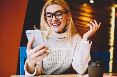 Neşeli hippi kız akıllı telefondan gelen iyi haberlerle heyecanlanıyor kafeteryada oturuyor, duygusal kadın internet iletişimi için indirimden memnun. Haberleri telefonla kontrol ediyor.