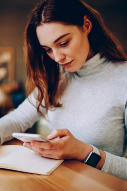 Ciddi kadın serbest çalışan not defterinde cep telefonuyla iletişim kuran, birlikte çalışma alanında çalışan, teknolojiyi ve bilgi aramak için kablosuz bağlantıyı kullanan genç bir kadın.