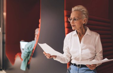 Beyaz gömlekli kıdemli Kafkasyalı iş kadını odaklı belgeleri inceliyor, ergonomik kırmızı koltuklu modern bir ofiste duruyor, aktif bir ilişki ve profesyonelliği öneriyor.
