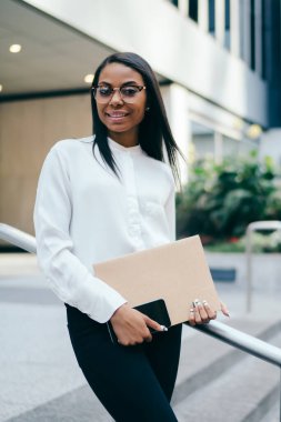 Mutlu Afrikalı-Amerikalı kadın avukat portresi kamera ayakta mimari ofis kentsel ortamda Binası yakınındaki gülüyorsun beyaz gömlek giymiş. Başarılı koyu tenli çalışan klasörü