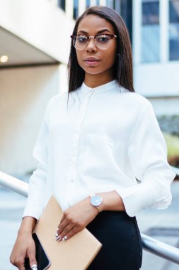 Beyaz tişörtlü başarılı Afro-Amerikan kadın çalışanın yarım boy portresi. Elinde akıllı telefon ve klasörle ofis binasının yanında kameraya gülümserken elinde belgeler var.