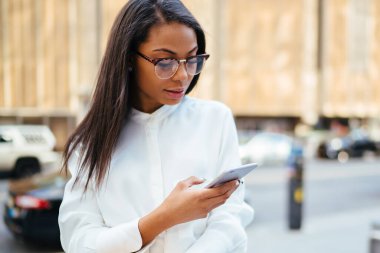 Afrikalı-Amerikalı kadın işçiye giyinmiş şık gömlek 4 g ile bildirim gönderme smartphone aygıt ayakta şehir merkezinde kentsel ortamda internette. Posta kontrol koyu tenli ofis çalışanı