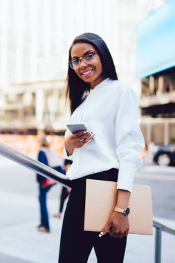 Klasör çevrimiçi smartphone aygıt ayakta şehir merkezinde sohbet elinde başarılı koyu tenli avukatla tam uzunlukta portresi. Müreffeh Afrikalı-Amerikalı genç kadın üzerinde cep SMS gönderme