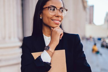 Şık formal giyim uzağa bakarak düşünceli gülümseyen Afrikalı-Amerikalı kadın işçiye. Koyu tenli klasör ve smartphone kentsel ortamda ayakta elinde tutan durulması ofis çalışanı