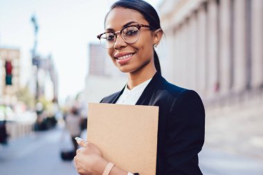 Kameraya gülümseyen elinde klasörle Hukuk Fakültesi başarılı Afro-Amerikan öğrenci portresi. Formal giyim açık havada duran neşeli kadın memur koyu tenli giyinmiş