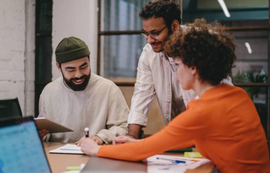 Yoğunlaşmış sakallı erkekler ve günlük giysiler ve gözlüklü kıvırcık saçlı bayan meslektaşlar laptopla masaya oturup yaratıcı çalışma alanı projesinde çalışırken notlar alıyorlar.